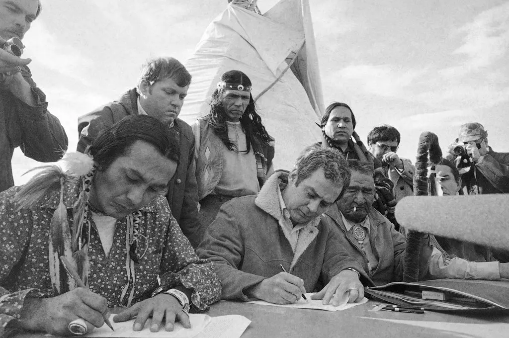 Dennis Banks, com faixa na parte traseira, observou seu colega líder do Movimento Indígena Americano, Russell Means, na frente à esquerda, assinar um acordo com o procurador-geral assistente Kent Frizzell em Wounded Knee, SD, em 1973.Crédito...Imprensa Associada