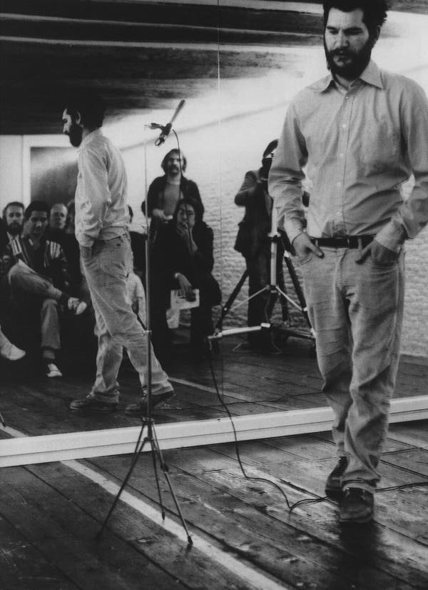 O Sr. Dan Graham em uma performance de 1977. De pé, entre uma parede espelhada e uma plateia sentada, ele os instruiu sobre o que estavam observando.Crédito...Dan Graham, através da Galeria Lisson